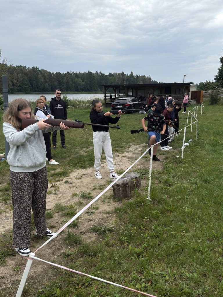 Młodzież uczestniczy w zajęciach strzeleckich na świeżym powietrzu, ćwicząc celność z broni sportowej.