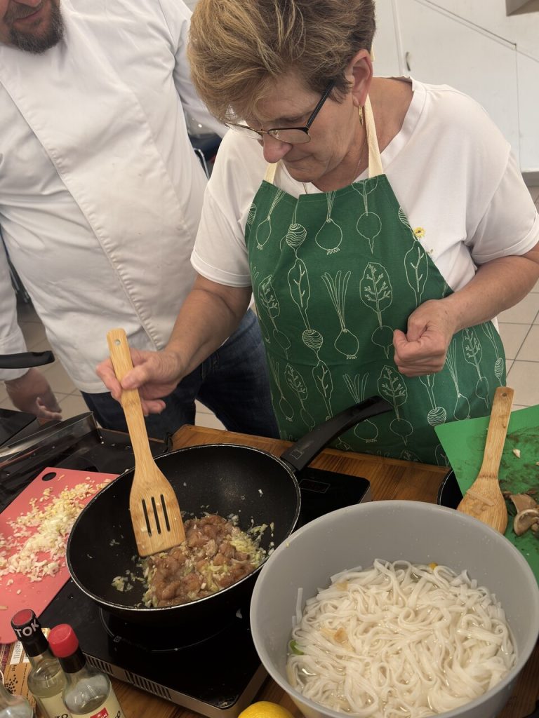 Kobieta w zielonym fartuchu gotuje potrawę na patelni, obok mężczyzna i składniki na stole.