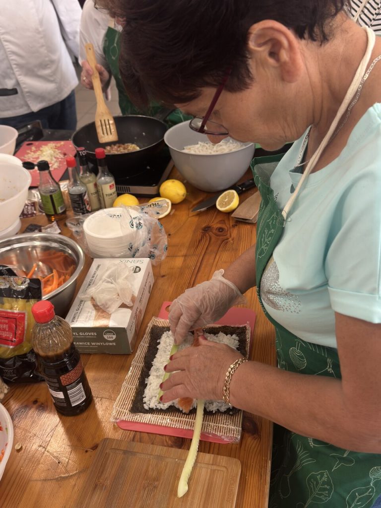Kobieta w zielonym fartuchu przygotowuje sushi, układając składniki na macie do zwijania.