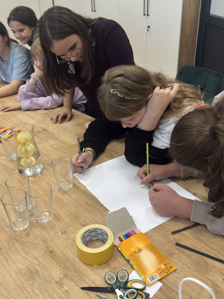 Dzieci rysują na dużym arkuszu papieru, otoczone przyborami plastycznymi i szklankami z wodą z cytryną.