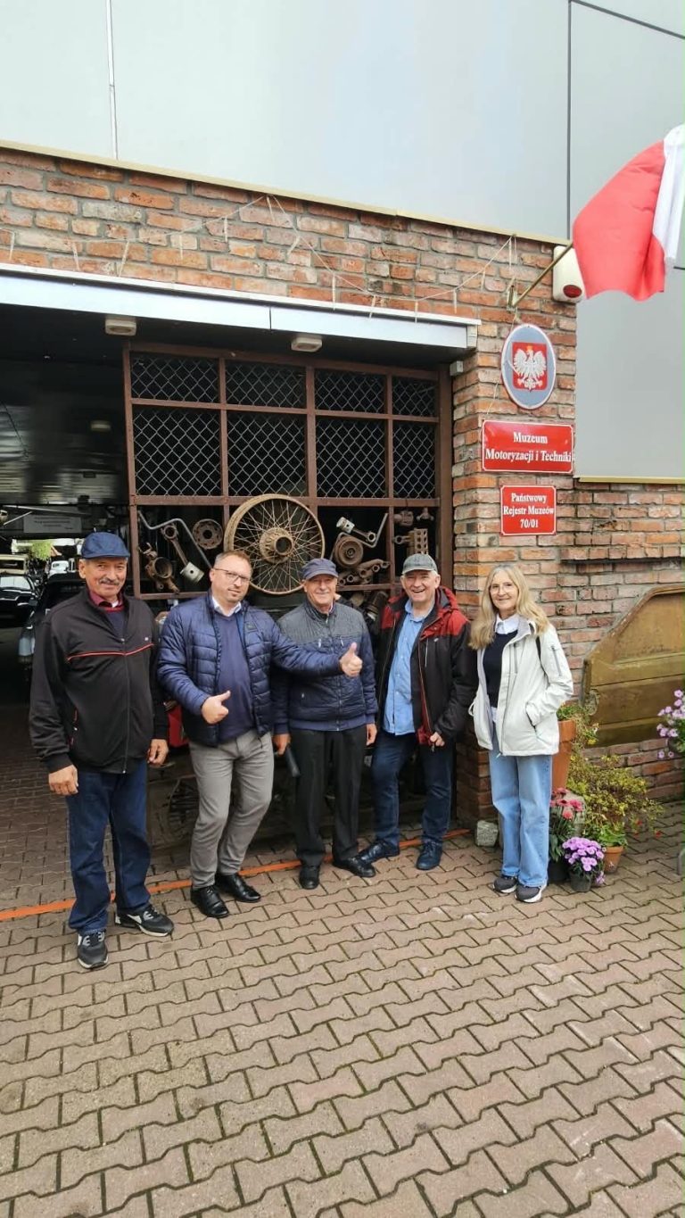 Grupa osób stoi przed Muzeum Motoryzacji i Techniki, otoczona eksponatami motoryzacyjnymi i flagą Polski.