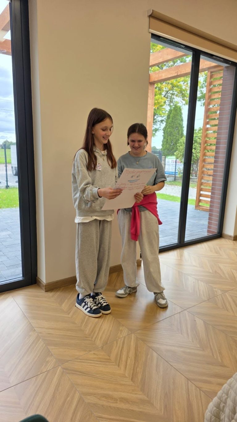 Dwie dziewczynki w szarych dresach czytają dokument w jasnym wnętrzu z dużymi oknami.