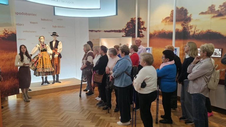 Grupa osób słucha przewodnika objaśniającego tradycyjne stroje ludowe w muzeum.