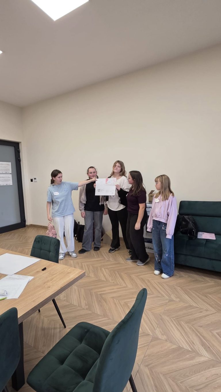 Grupa osób prezentuje plakat w jasnym pomieszczeniu, z drewnianym stołem i zieloną kanapą w tle.