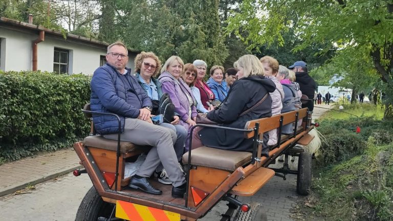 Grupa osób siedzących na wozie, ciesząca się wspólną przejażdżką w otoczeniu zieleni.