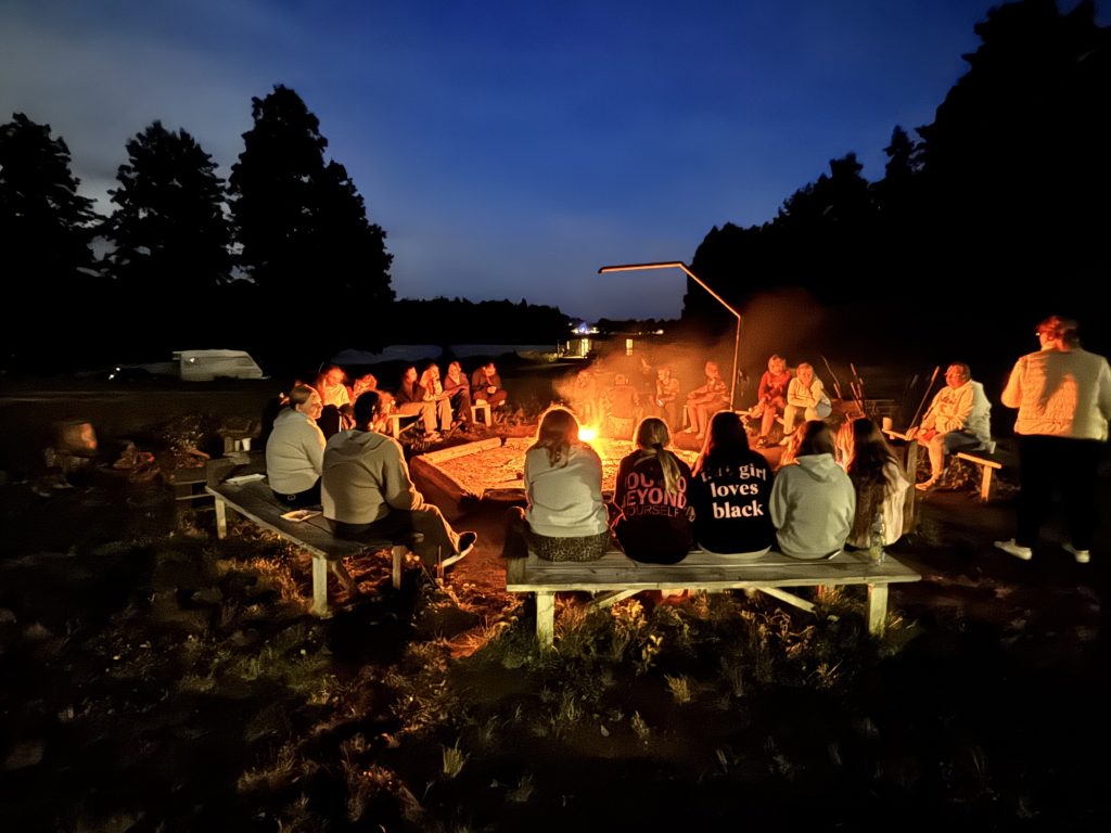 Grupa ludzi siedzi wokół ogniska w nocy, ciesząc się wspólnym czasem i atmosferą.