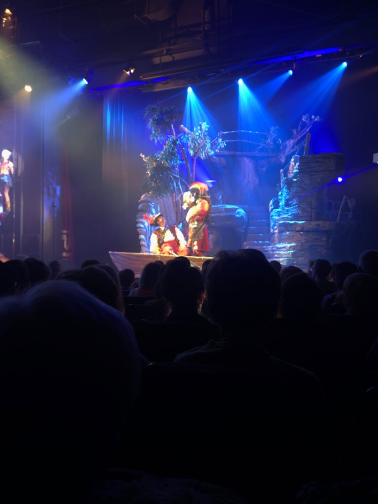 Występ teatralny z piratami na scenie, oświetlony kolorowymi światłami, z widownią w tle.