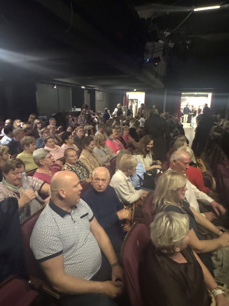 Widownia teatru z tłumem ludzi siedzących na czerwonych fotelach, czekających na rozpoczęcie spektaklu.